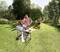 A group of four people are barbecuing outdoors on a verdant lawn. Two people are standing by the barbecue and appear content, while another person is stoking the barbecue with charcoal.