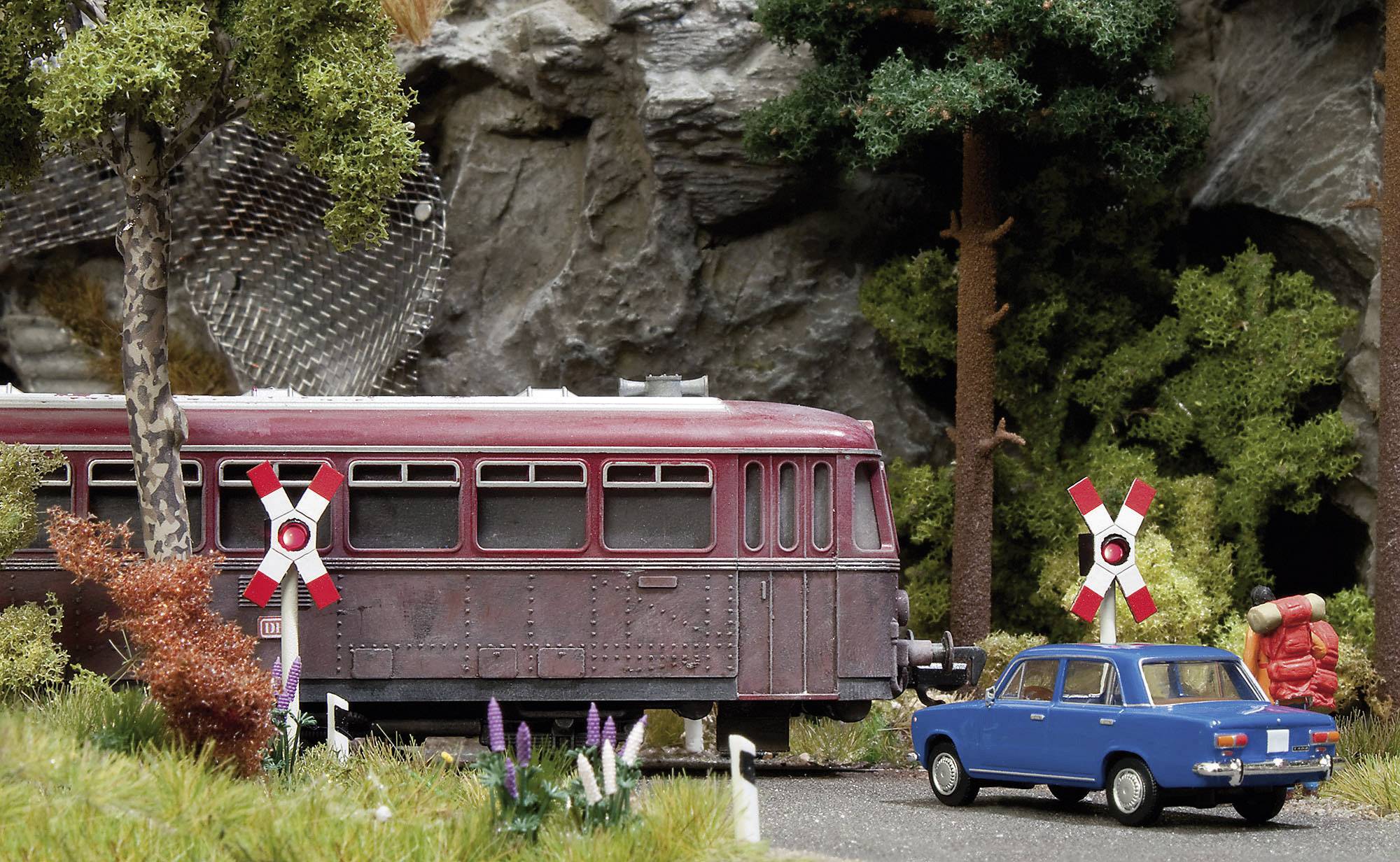 A blue car is waiting at a level crossing while a red train with a cross symbol passes by. Forest and rocks are in the background.
