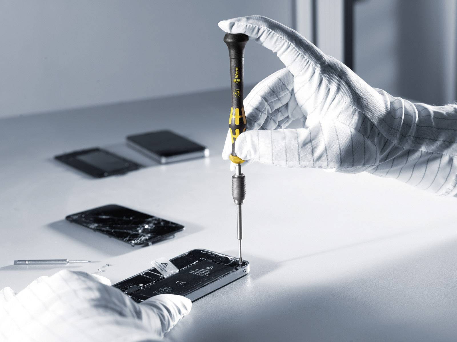 A person wearing gloves is repairing a smartphone with a screwdriver. Additional smartphones and tools are lying on the table.