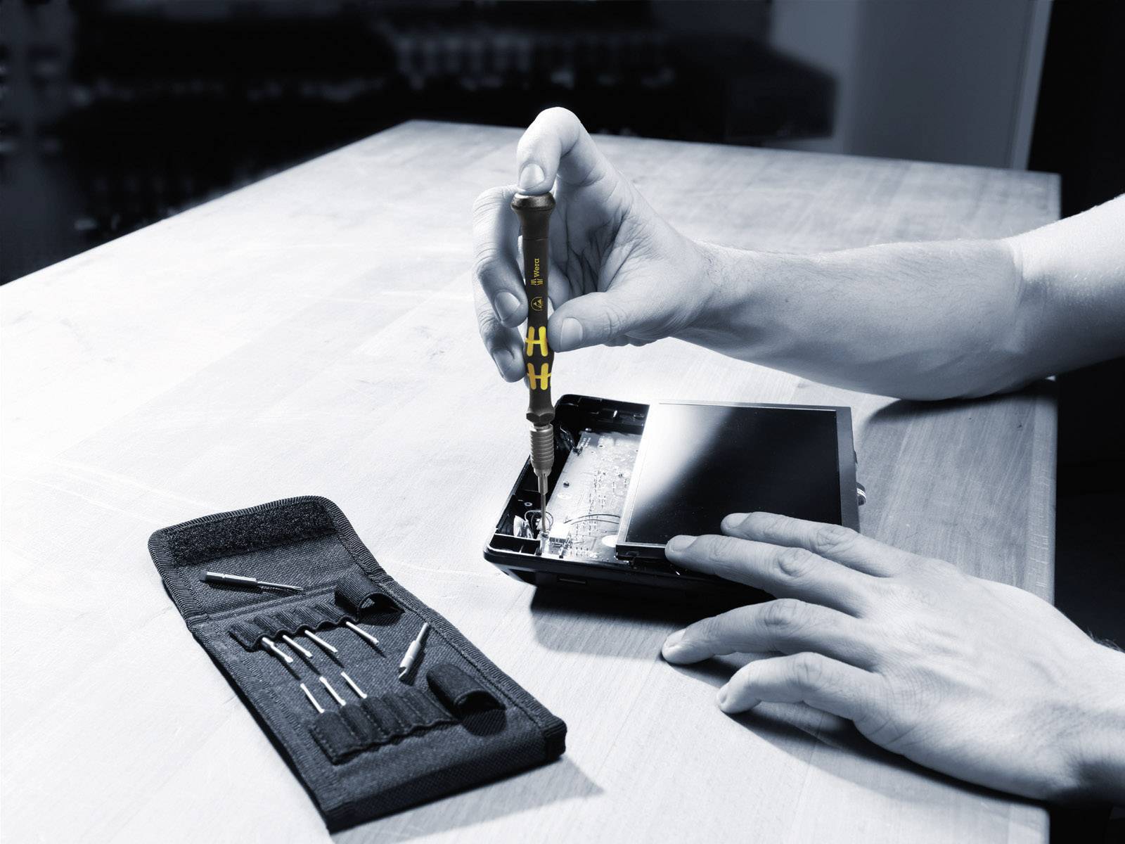 A person is repairing a device with a screwdriver. A set of screwdriver bits in a case lies on the table.