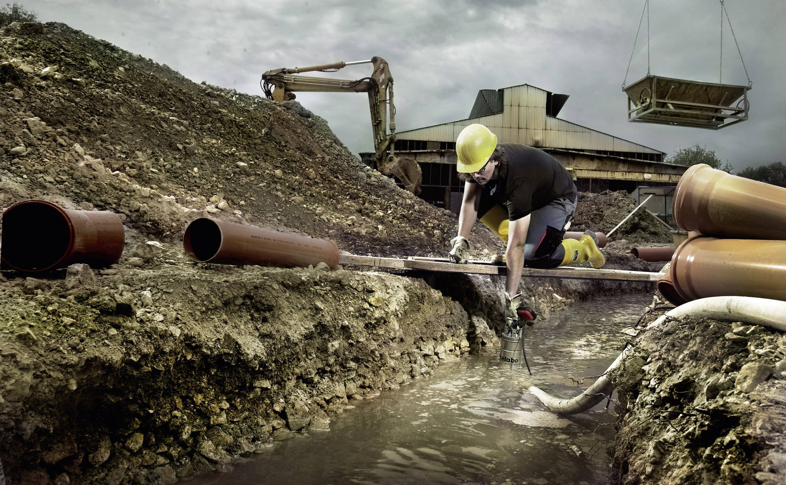 A construction worker wearing a hard hat kneels in a waterlogged trench, working on a pipe. In the background, an excavator and a building are visible.