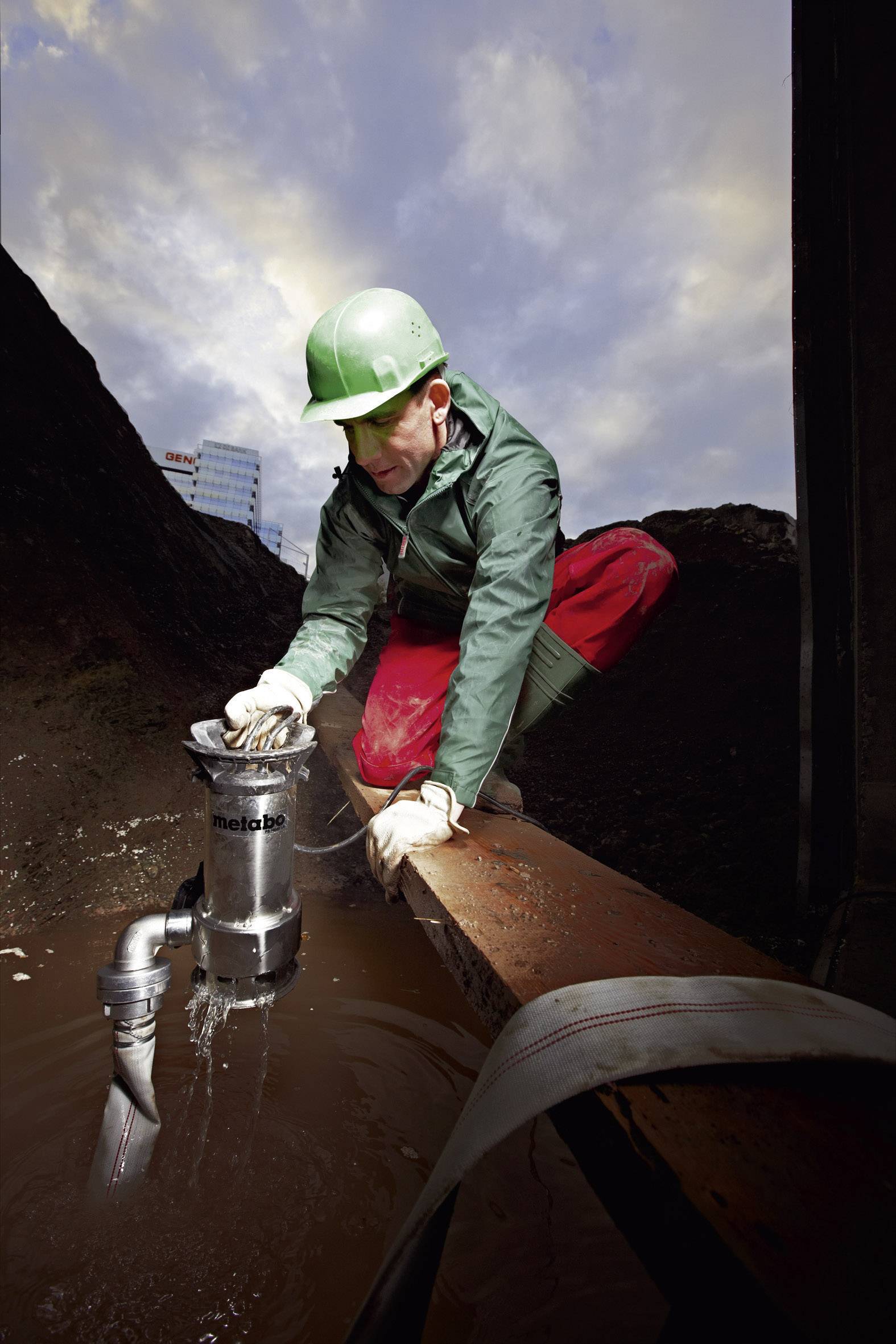 A construction worker wearing a green hard hat is installing a pump in a muddy area on a building site under a cloudy sky.