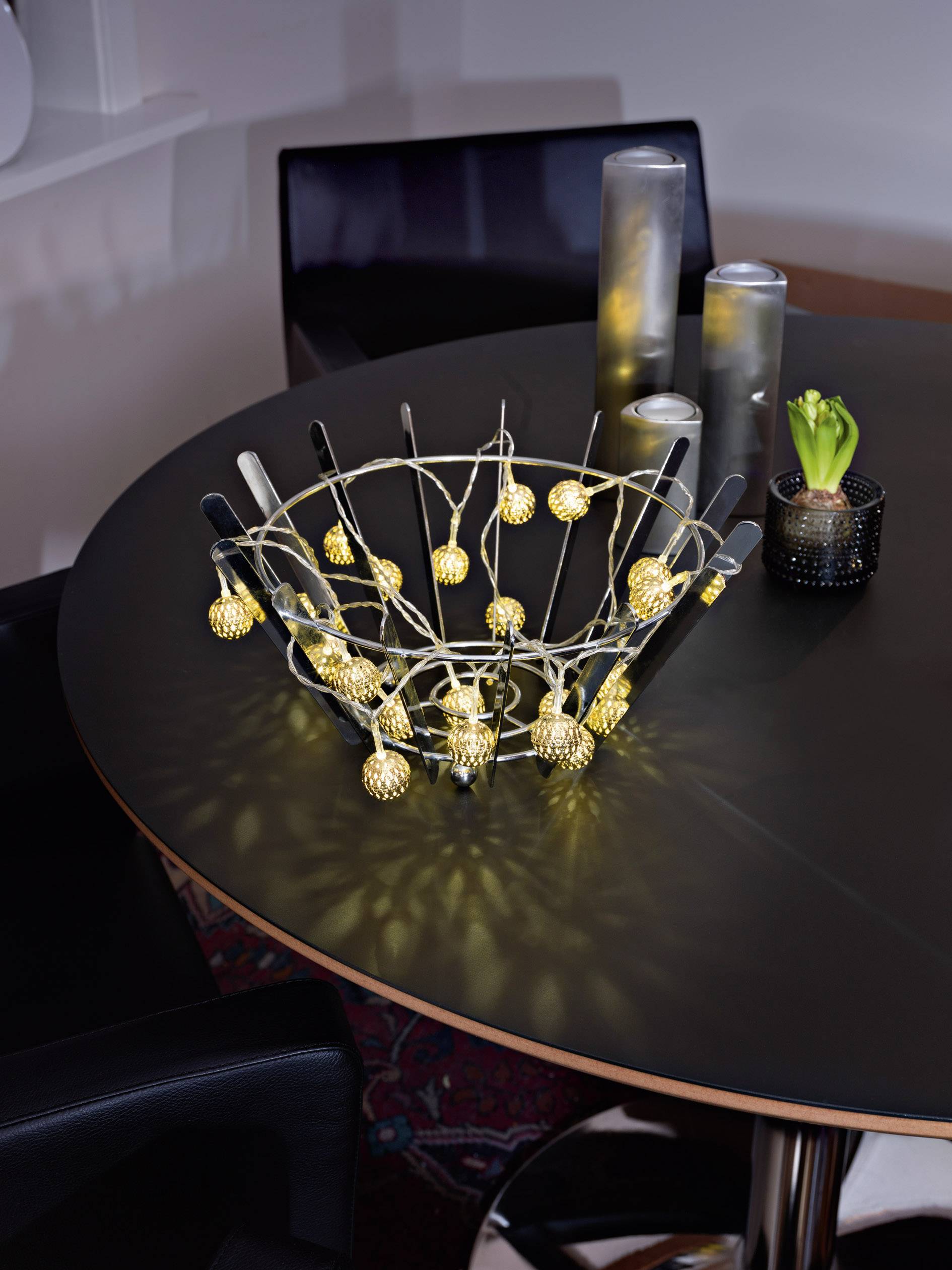 A round table with decorative lights in a basket in the centre, accompanied by candles and a small green plant.