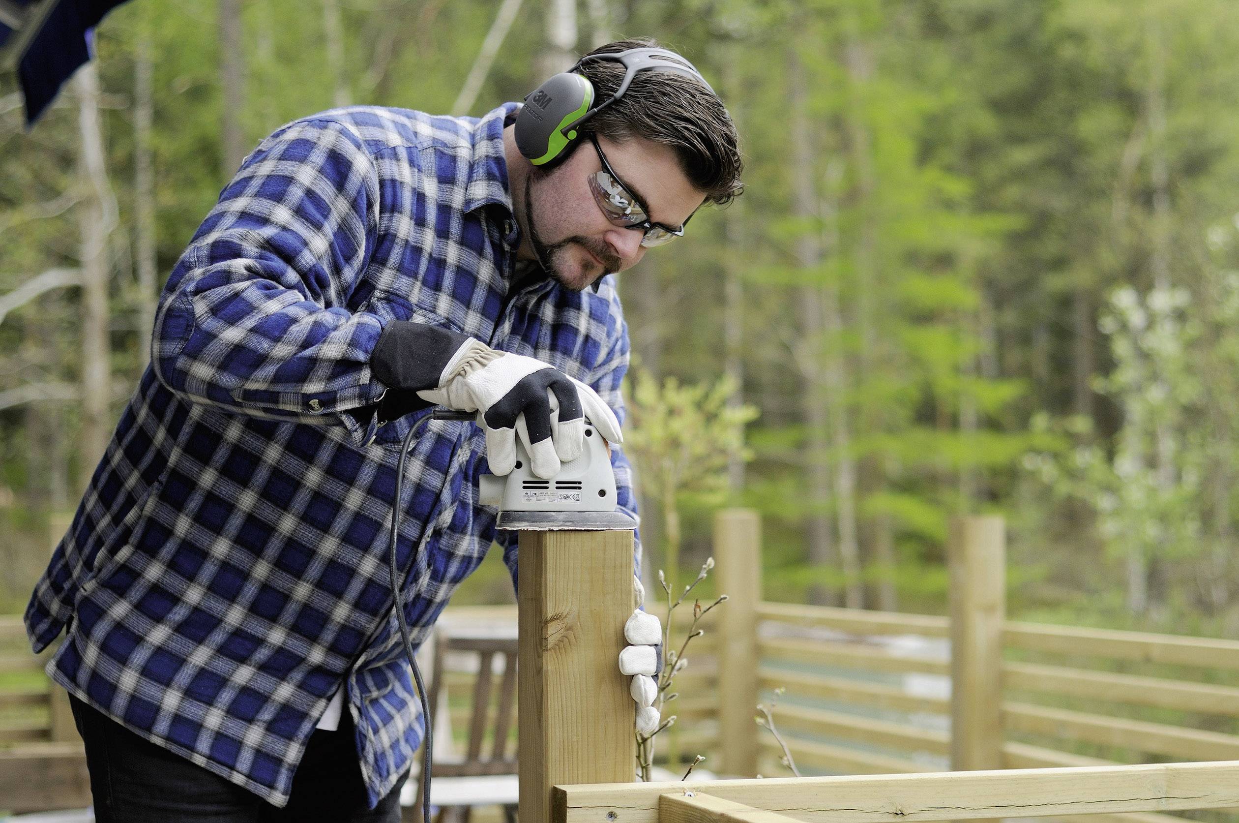 A man is working with an electric sanding machine on a wooden terrace, wearing safety glasses and ear protection, surrounded by woodland.