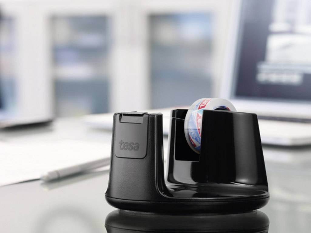 A black tape dispenser with the inscription 'tasa' sits on a desk in front of a blurred laptop background.