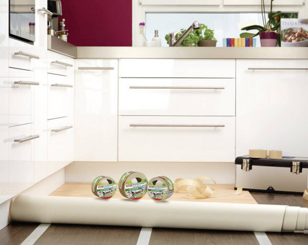 White kitchen with open storage. Vinyl flooring roll, adhesive tape, and tiling tools scattered on the floor. Plants on the worktop.