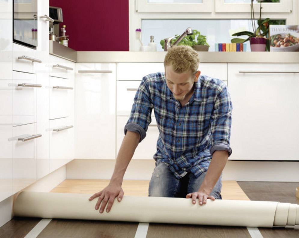 A man is laying a new floor covering in a modern kitchen. The kitchen is bright, with white cupboards and plants in the background.
