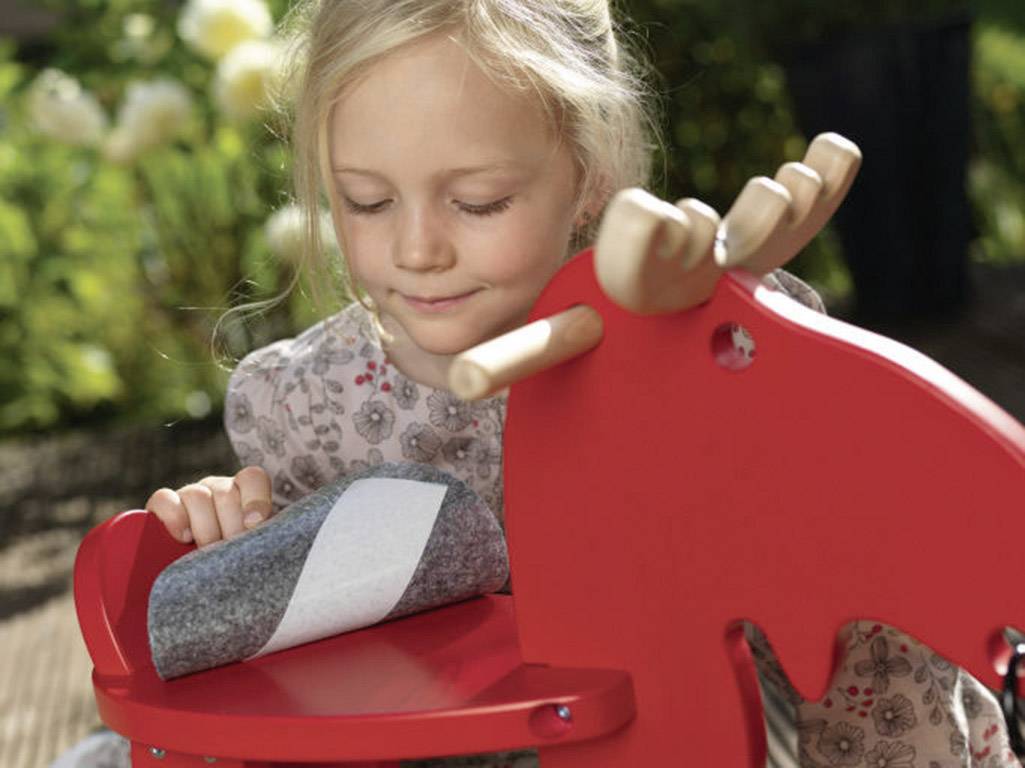 A child is playing outside with a red wooden toy elk. The child appears happy and focused. Flowers are blurred in the background.