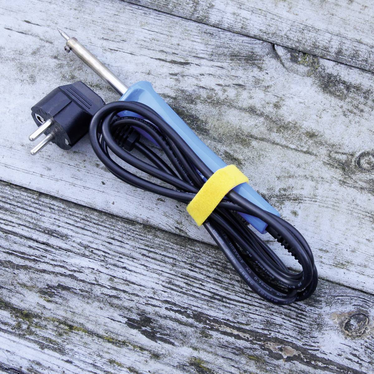 A soldering iron with a blue handle and European plug is resting on a rustic wooden table.