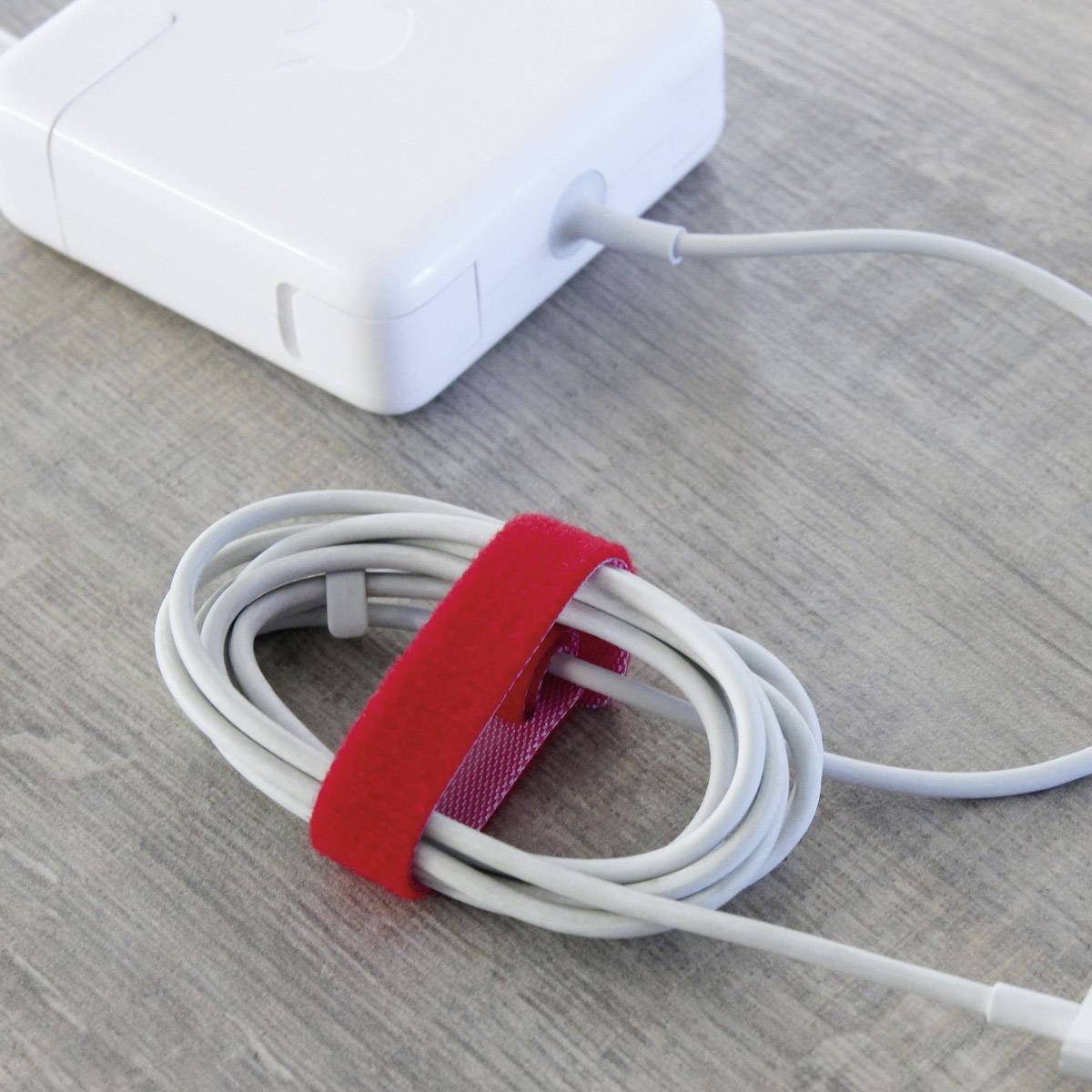 A white laptop power adapter is sitting on a table, beside it a coiled cable secured with a red hook and loop fastener.