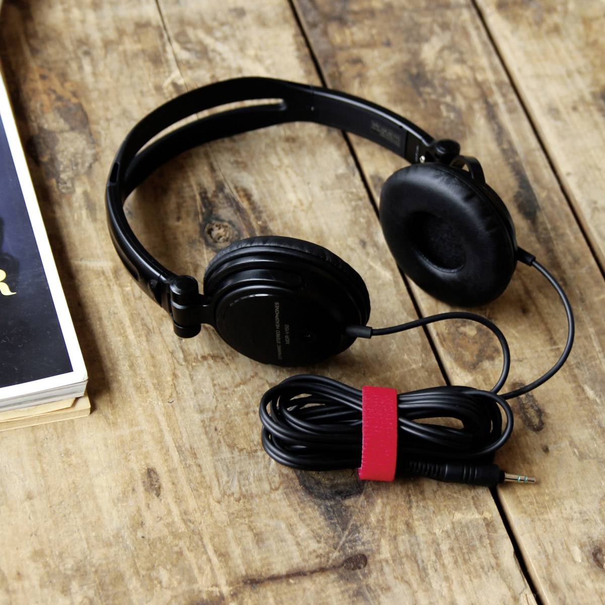 A black pair of headphones with a coiled cable lies on a wooden table next to a stack of books.