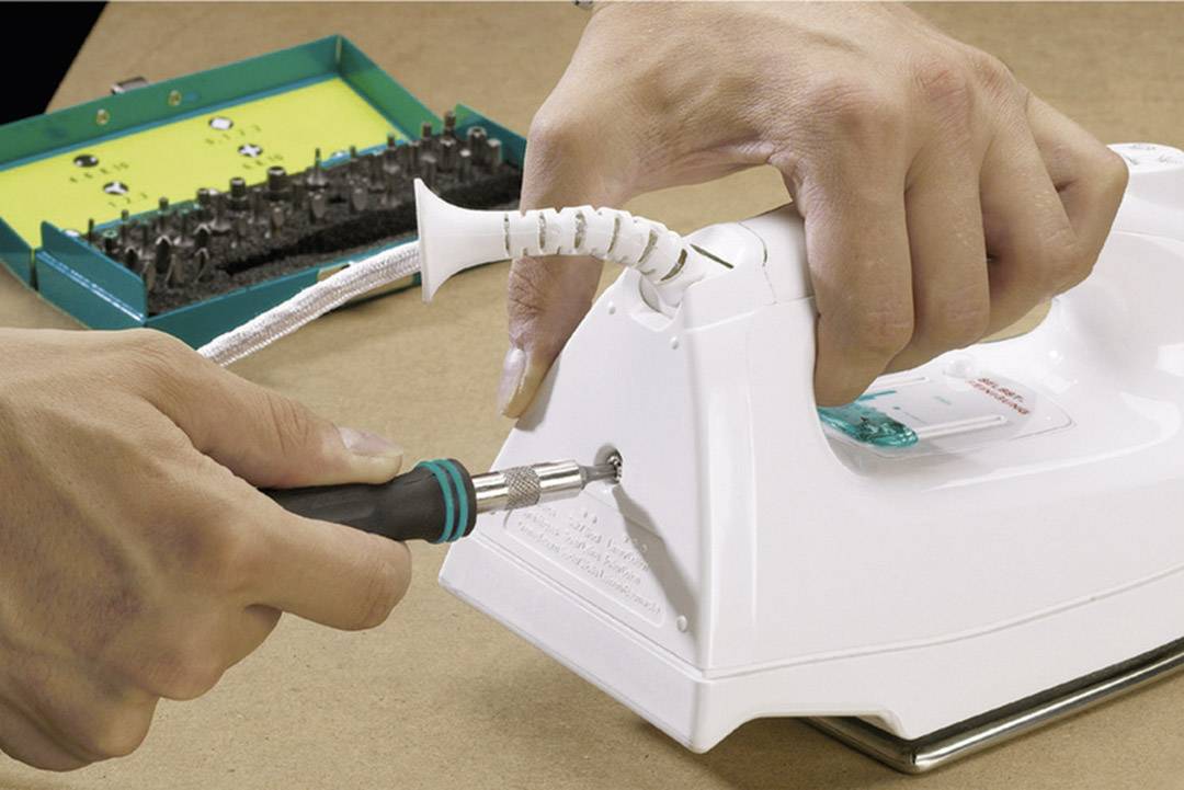 A person is repairing an iron by loosening a screw with a screwdriver. A tool set is visible in the background.