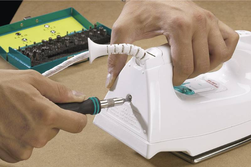 'Person repairs iron with screwdriver', shows hands tightening a screw on a white iron. Toolbox in the background.