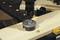 A cordless drill is drilling a hole in a wooden board, secured on a workbench. Wood shavings are visible around the drilling area.