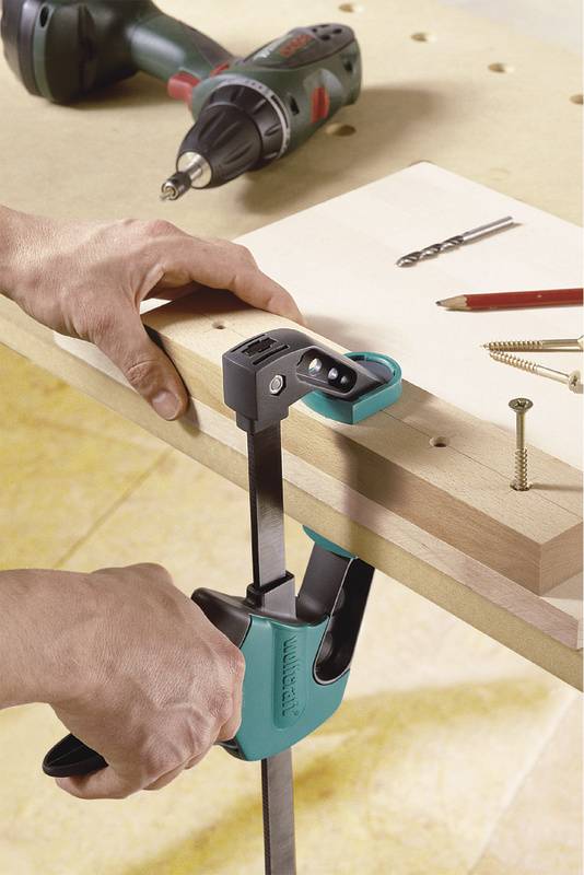 A person is securing a wooden board to a table with a clamp. A drill, pencil, and screws are lying on the table.