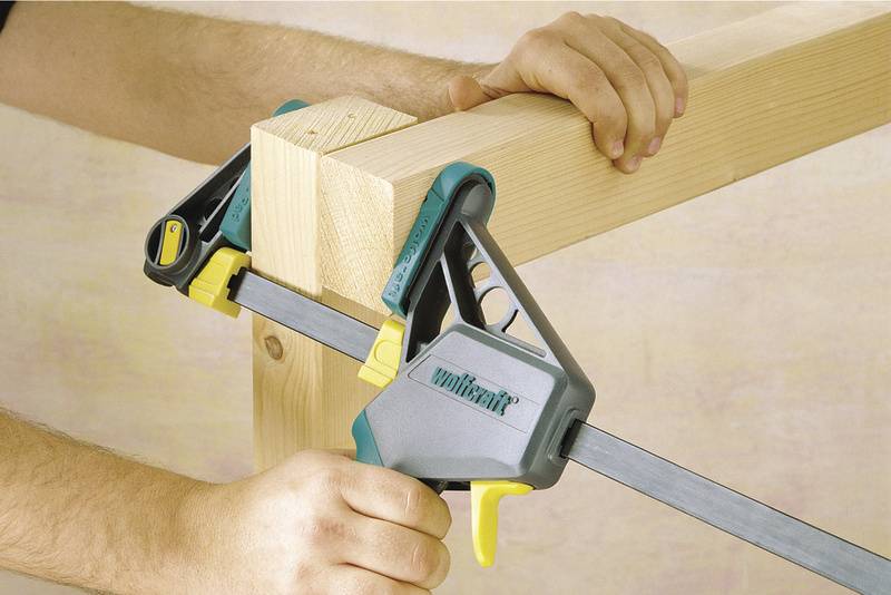 A person is using a one-handed clamp to hold two pieces of wood at right angles and firmly together.