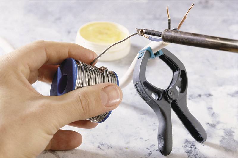 A hand holds a solder reel while a soldering iron solders a cable. A clamp secures the cable on a white surface.