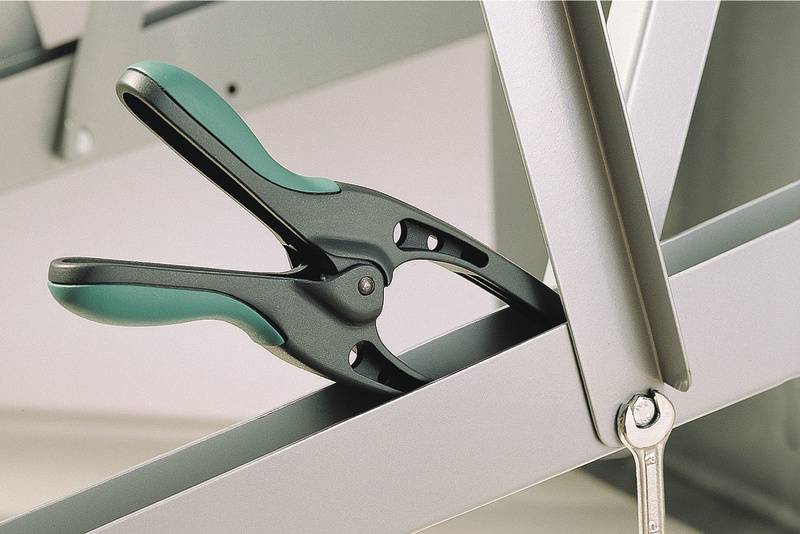 A black clamp with green handles holds two metal struts together. Underneath, a spanner hangs from the lower strut.