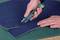 A person is cutting a blue carpet with a green Stanley knife on a cutting mat.