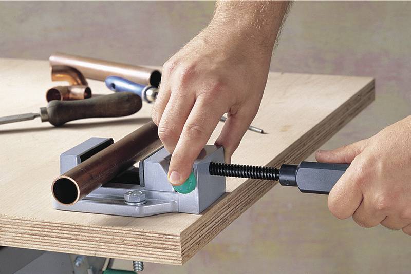 A person is holding a copper pipe with a G-clamp on a workbench, with various tools scattered around.