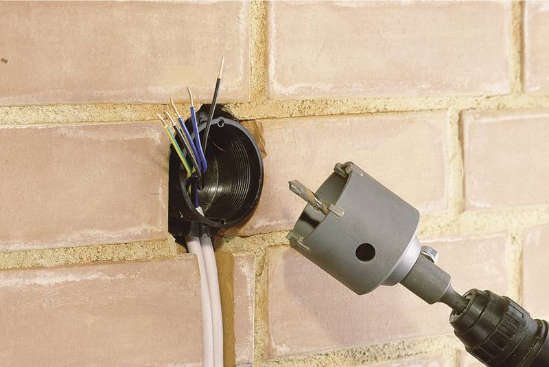 A drill with a hole saw is boring a circular hole in a brick wall. Cables are protruding from a round sleeve beside it.