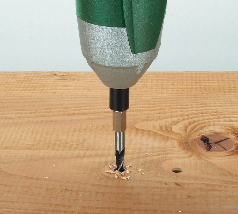 A green drill bit is drilling a hole in a wooden board, with wood shavings visible around the hole.
