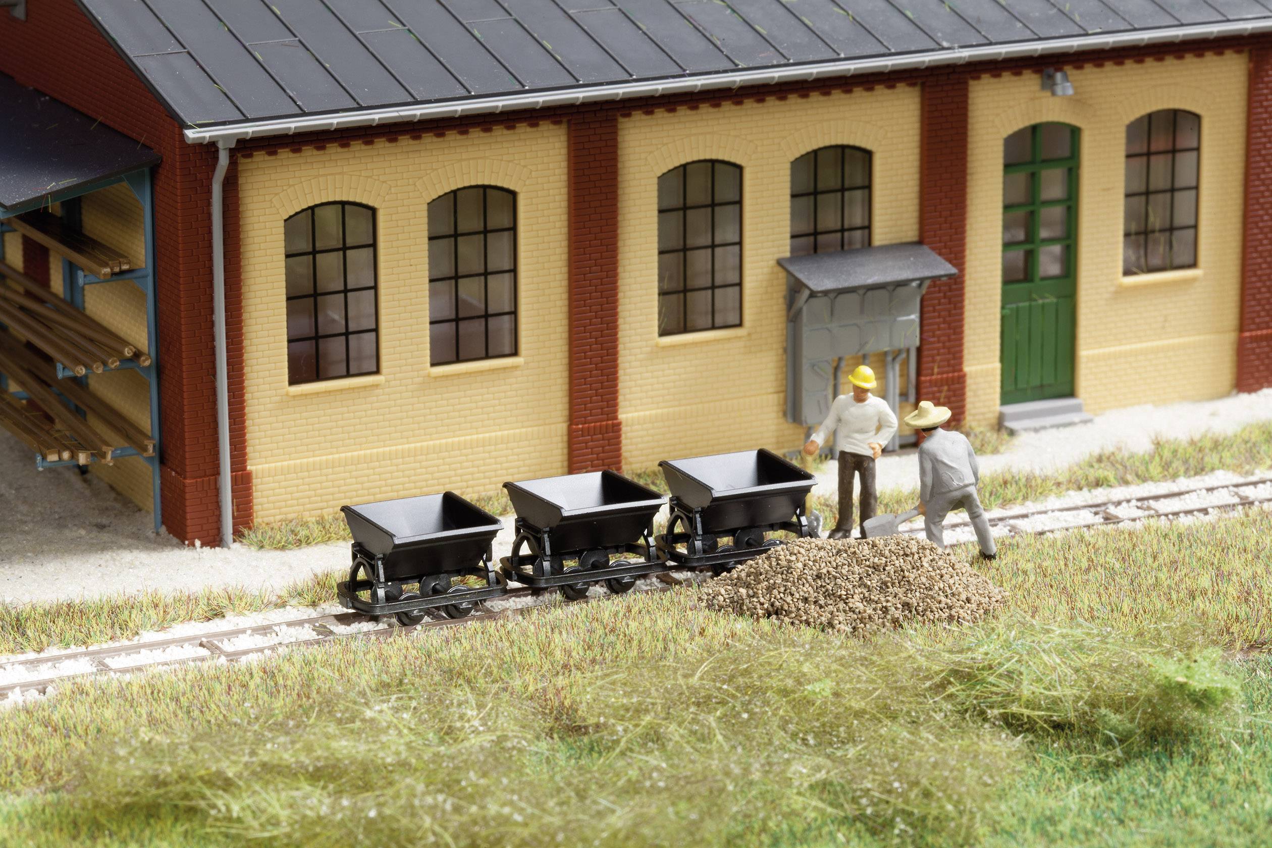 Two figures stand beside small wagons on tracks in front of a building. A pile of gravel lies nearby. It looks like a model railway scene.