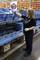 A person is sorting goods into blue crates on a conveyor belt in a warehouse. Stacked crates can be seen in the background.
