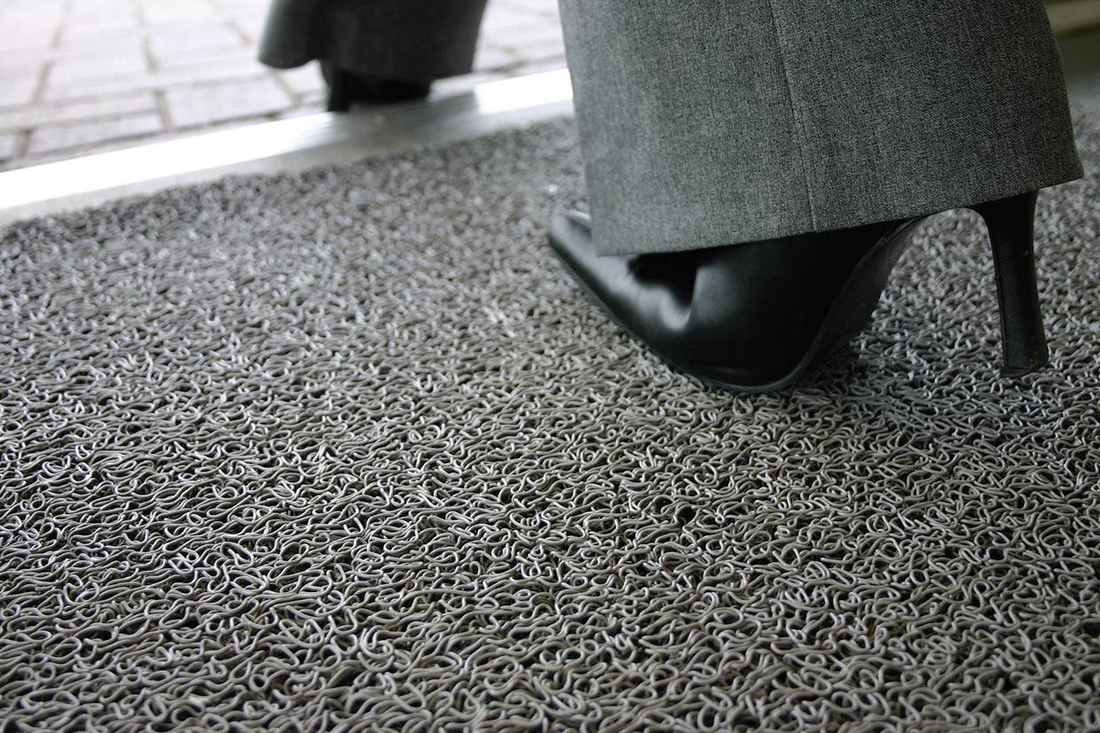 A black high-heeled shoe enters a building on a grey doormat.