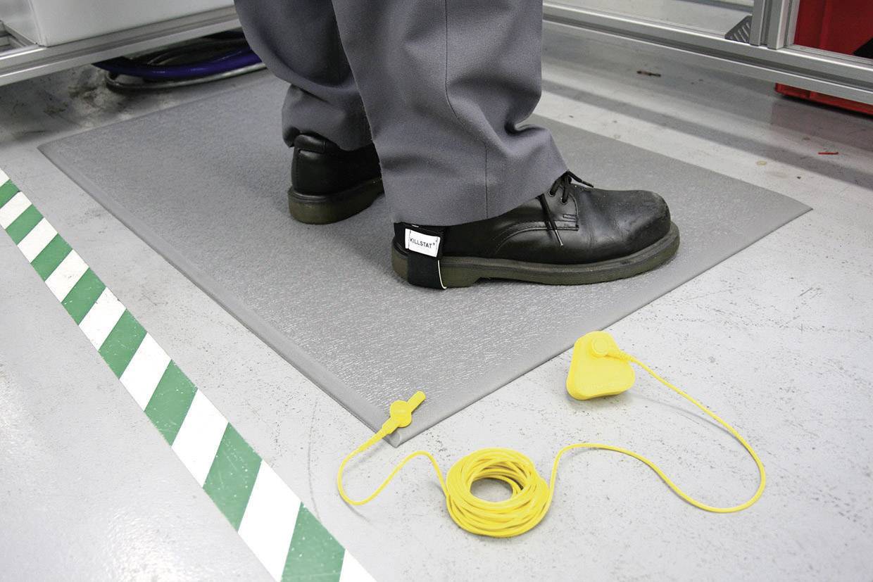 A standing person is wearing safety shoes on an anti-static mat. A yellow earthing cable lies on the ground beside them.