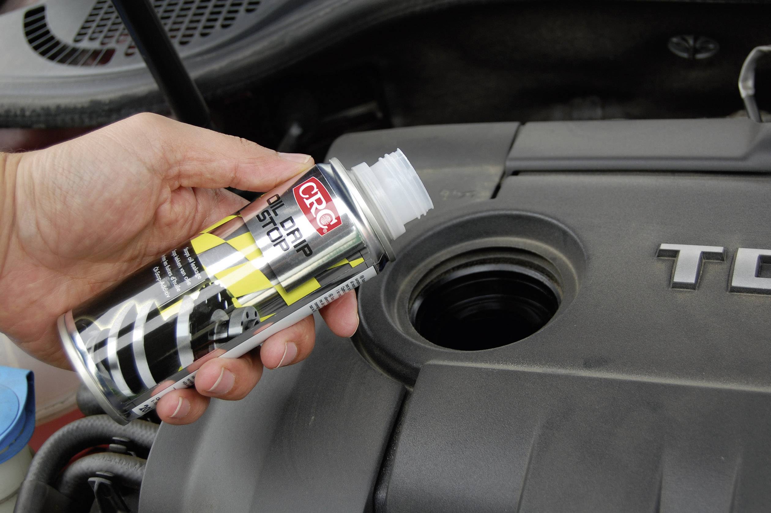 A person is pouring a liquid from a CRC can into the filler opening of a car engine, likely to improve performance.