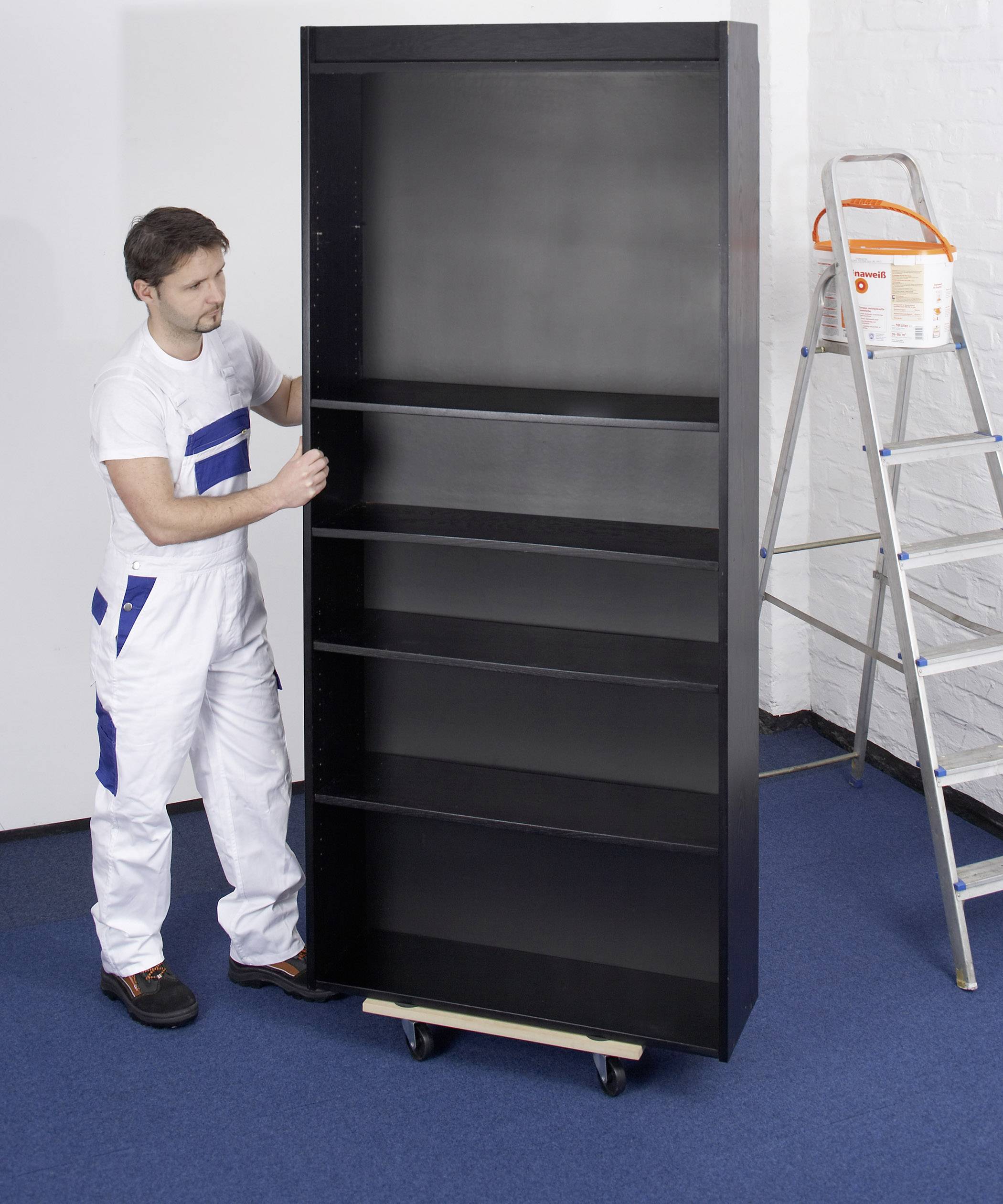 A man in workwear is positioning an empty, black shelving unit on a wheeled trolley in a room with blue carpet.