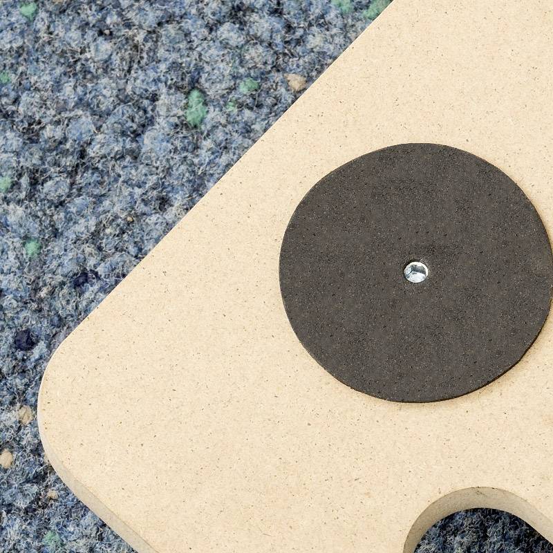 A round, black felt pad is fixed centrally to a light brown wooden board with a screw. The background is patterned carpet.