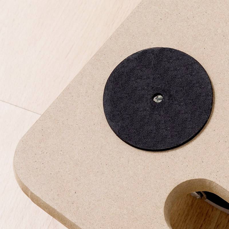 A rectangular wooden board with a round, black felt glider in the centre and a small screw on a light wooden floor.