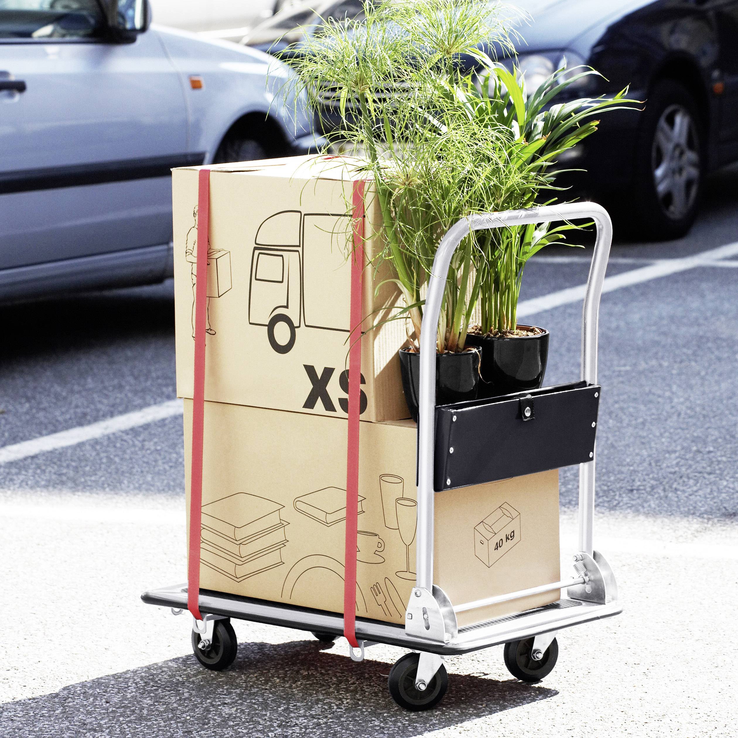 A trolley with cardboard boxes and plants is parked in a car park. The boxes are marked with icons and 'XS'.