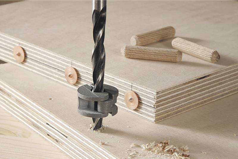 A drill with a depth stop is drilling into plywood boards. Wooden dowels for fastening are lying beside it.