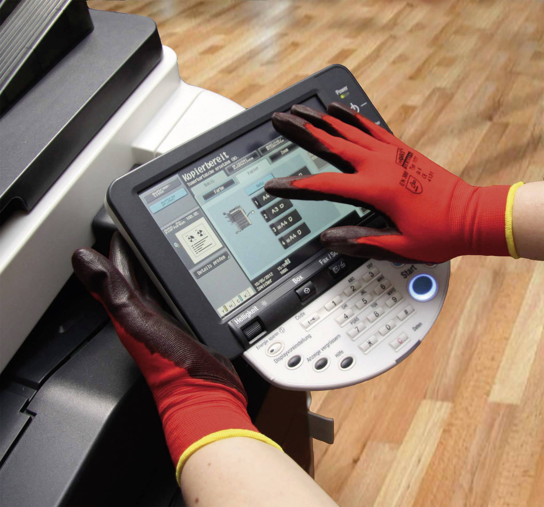 A person wearing red gloves is operating a printer control panel with a touchscreen. The screen displays printing options. A wooden floor is visible in the background.