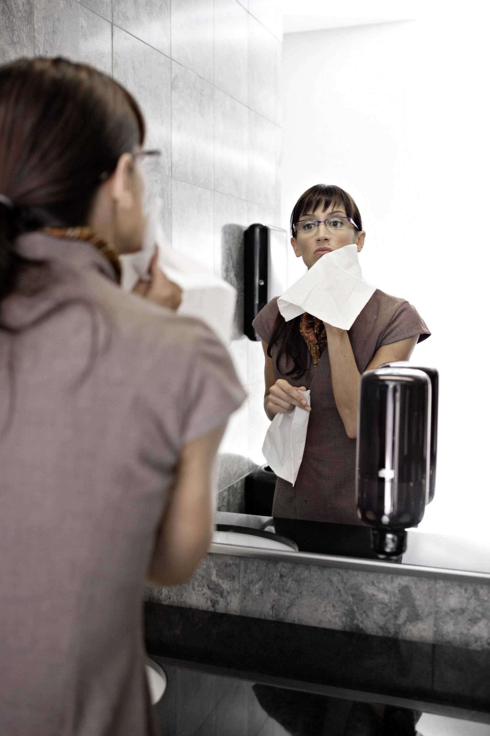 A woman stands in front of a mirror in a bathroom and dries her face with a paper towel.