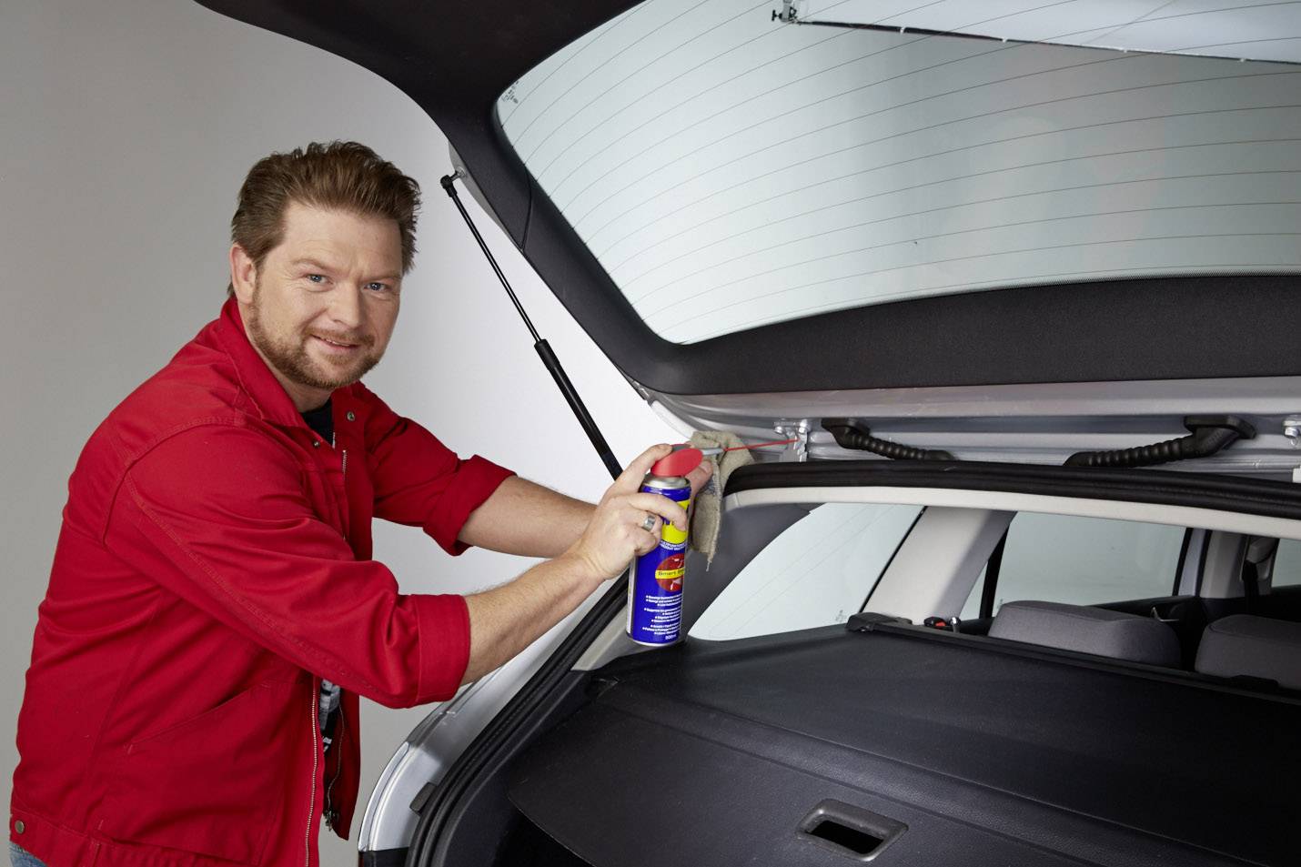A man in a red overall is spraying lubricant onto a car's boot lid.