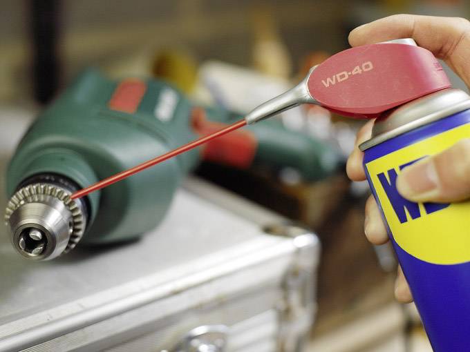 A hand sprays lubricant from a can with a long nozzle onto a green drill head to maintain or loosen it.