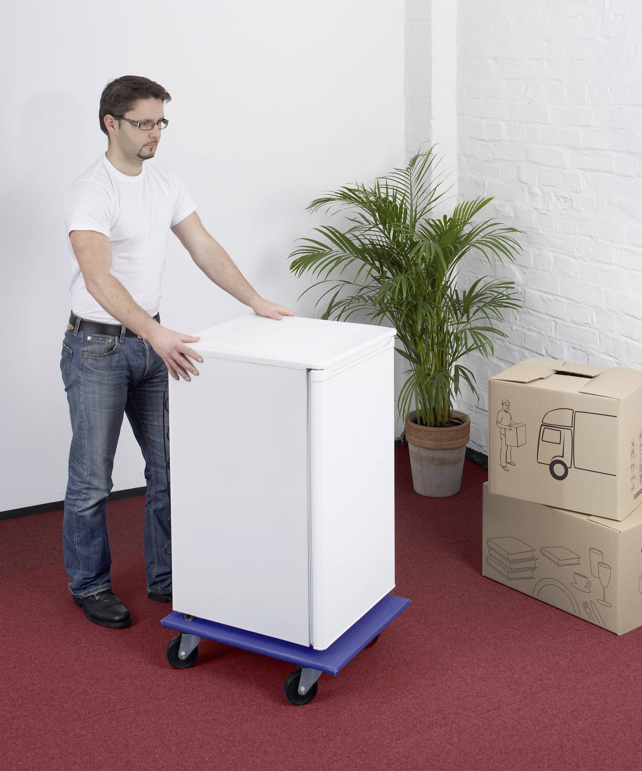 A man is moving a fridge on a blue trolley in a room with a red carpet and several cardboard boxes.