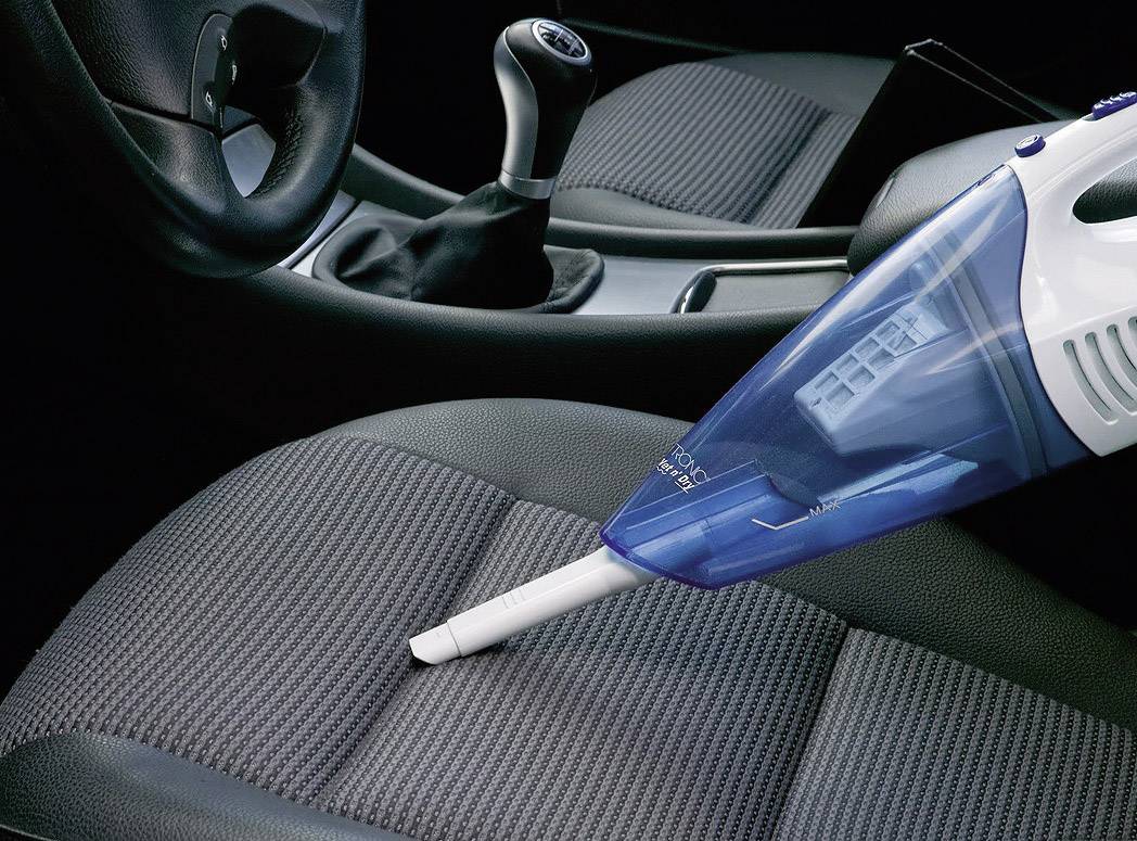 A handheld vacuum cleaner is cleaning a car seat. The dashboard and steering wheel are visible in the background.