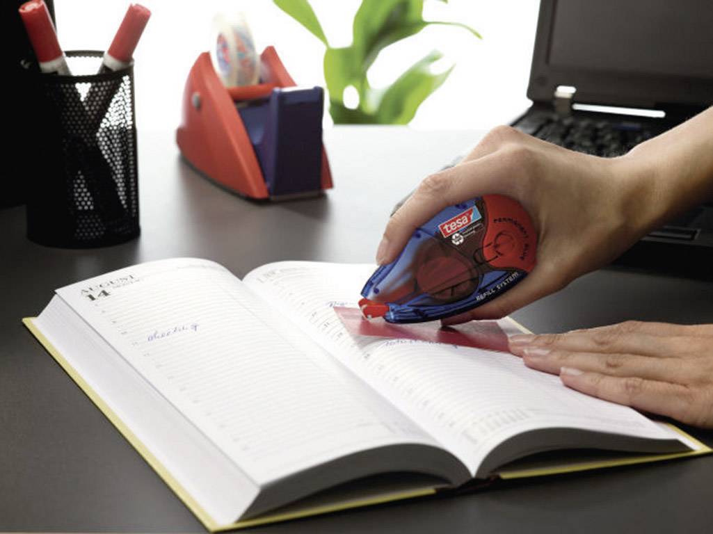 A person is using correction tape to amend text in an open notebook. Office supplies are visible in the background.