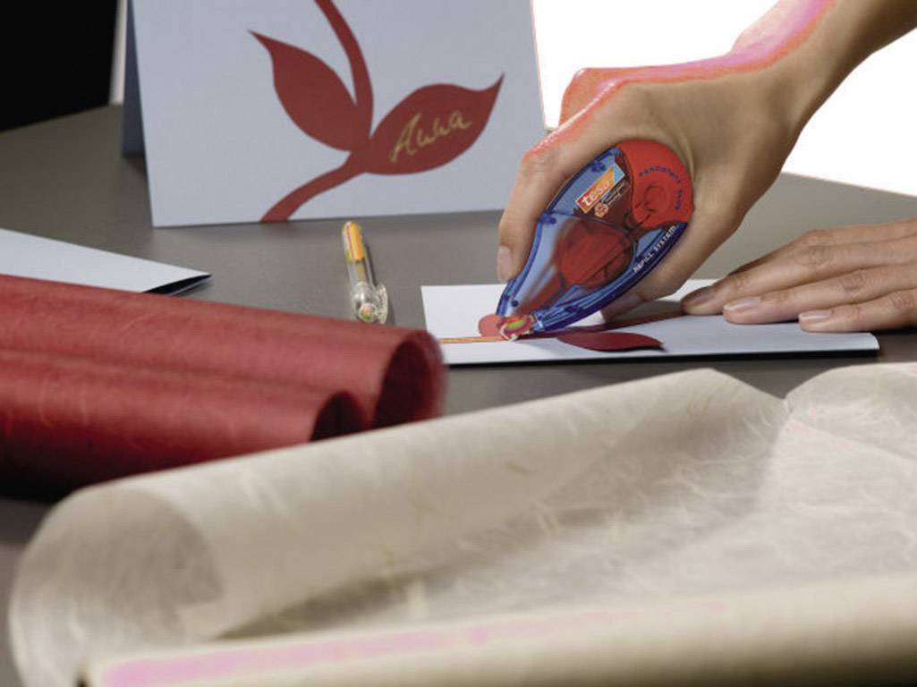 A hand is sticking a red leaf motif onto a greetings card using a glue roller. The name 'Alea' is written in red on the card.