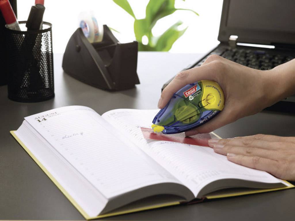 A person is correcting text in a notebook using a correction roller. A laptop and office supplies are visible in the background.