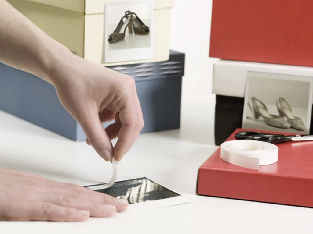 A hand is sticking a photo of shoes onto a box. Further shoeboxes with photos are visible in the background.