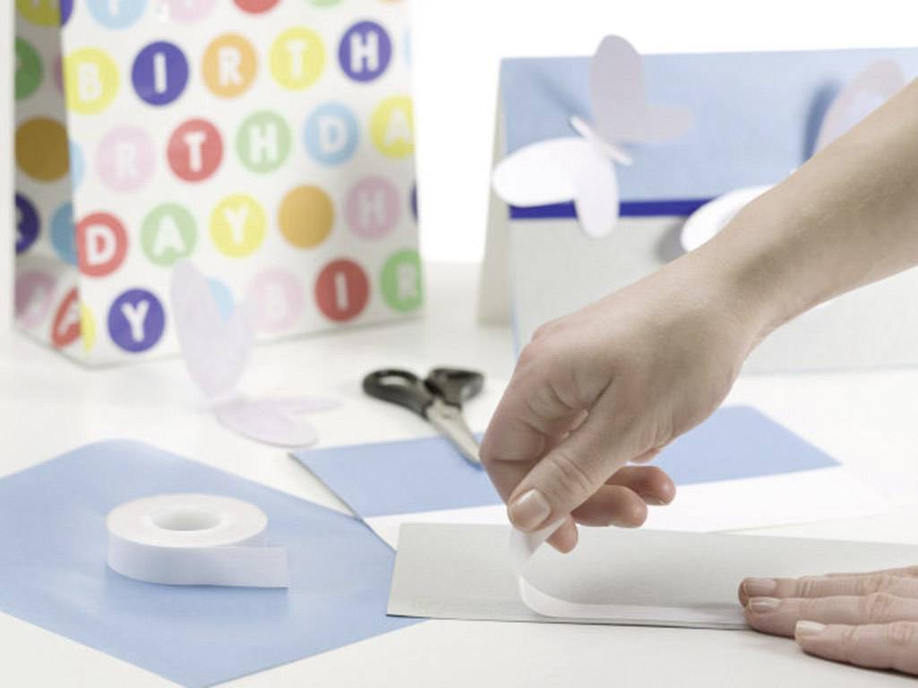 A hand is crafting with paper and sellotape on a table. In the background, a colourful birthday card and butterfly decorations are visible.