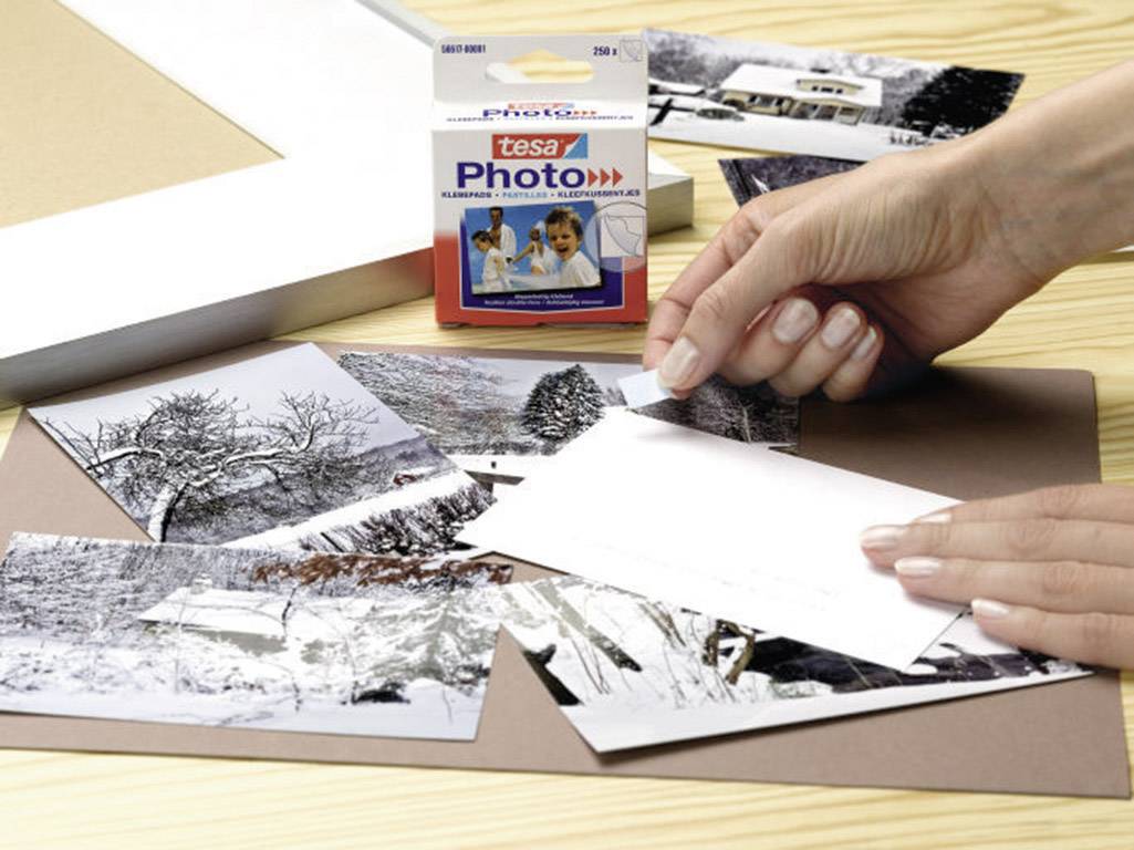 A person is sticking black and white photographs onto a surface using Tesa Photo adhesive strips. A pack of Tesa Photo strips is visible in the background.