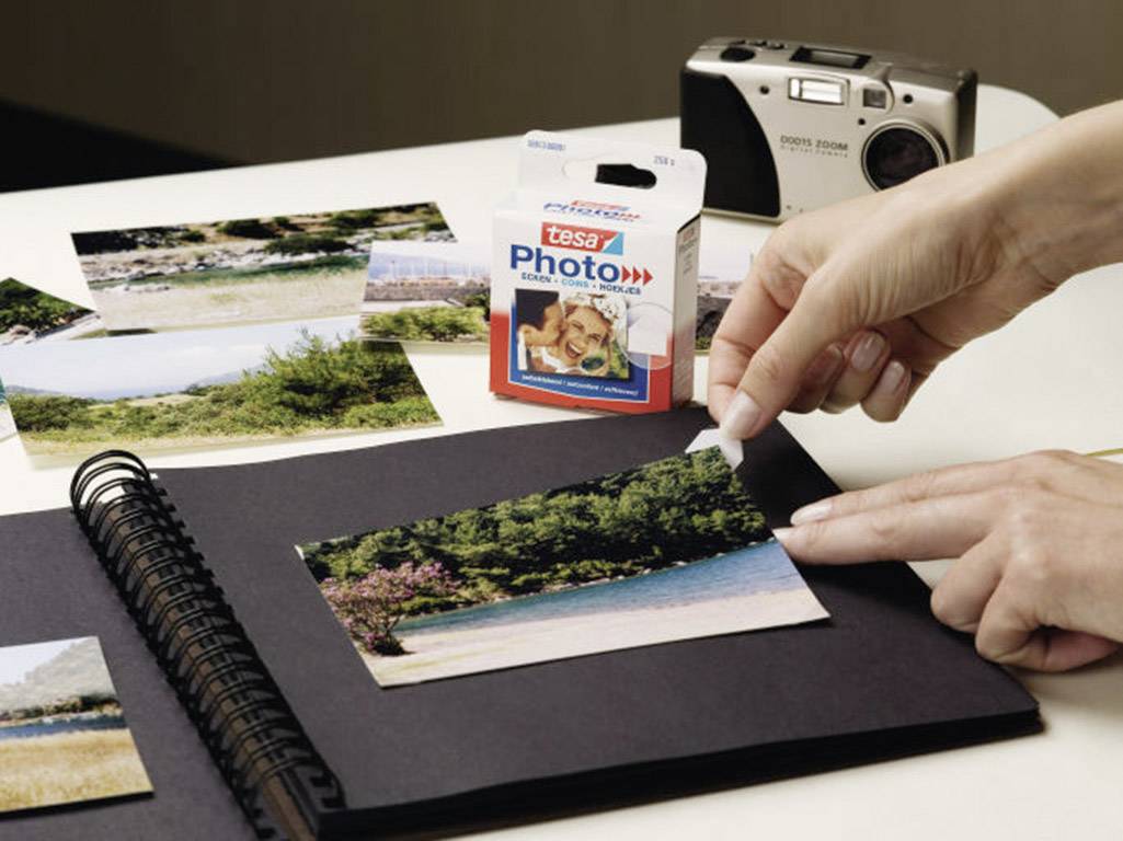 A person is sticking photos into an album using photo adhesive. A camera and additional photographs are visible in the background.