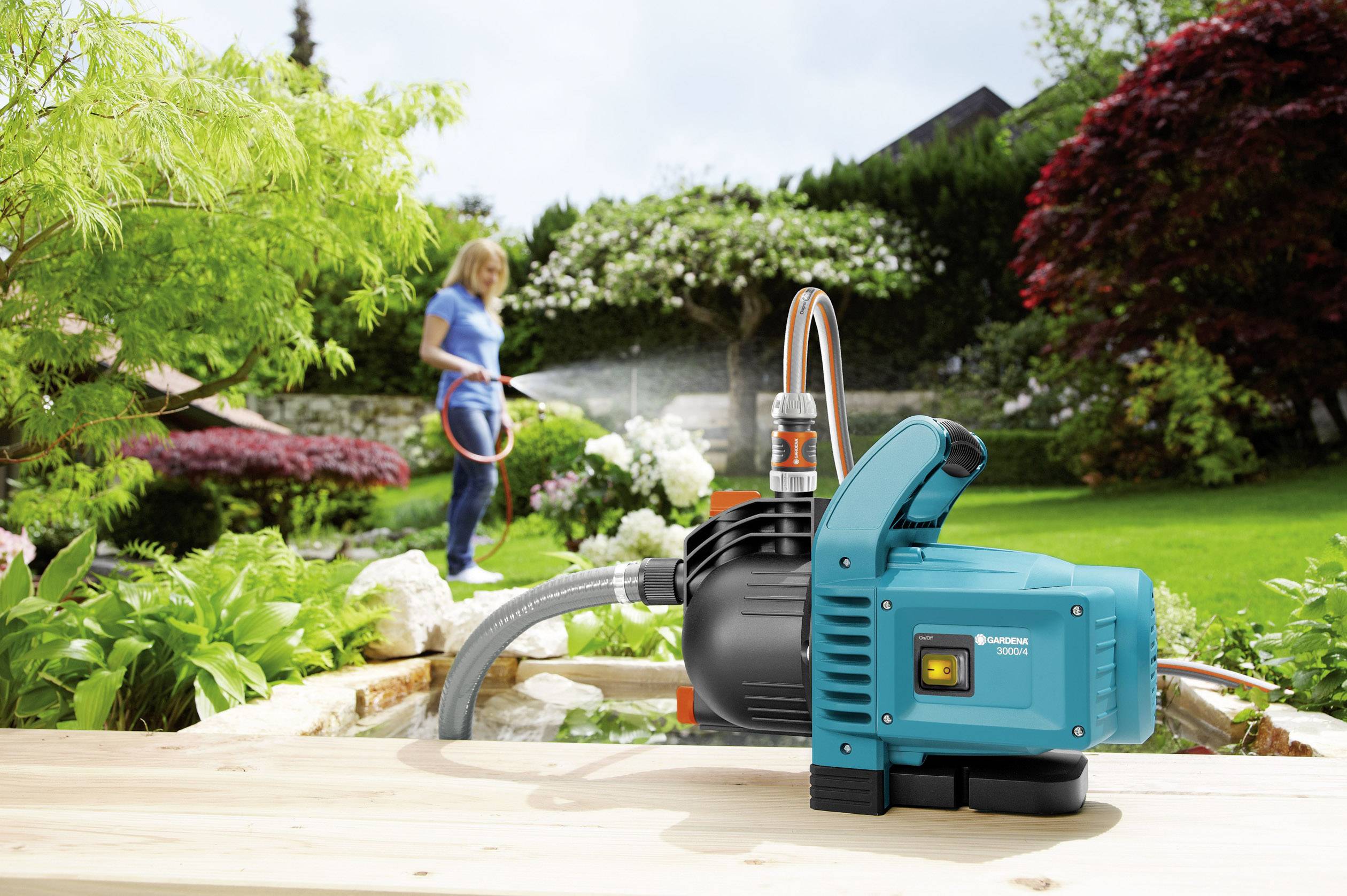 A person is watering plants in the garden using a garden hose. In the foreground, a garden pump is supplying the water.
